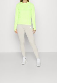 Kvinna i neon gul långärmad topp, beige Adidas leggings och vita sneakers står mot en enkel ljus bakgrund.