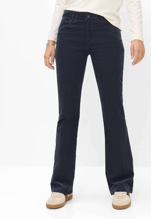 Frau trägt dunkelblaue ausgestellte Hose, weißes Langarmshirt, beige Sneaker, steht vor einem weißen einfarbigen Hintergrund.