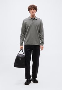 Νεαρός με σγουρά μαλλιά, φορώντας γκρι πουκάμισο πόλο με μακριά μανίκια, μαύρο παντελόνι, μαύρα παπούτσια, κρατώντας μια μαύρη δερμάτινη τσάντα ταξιδιού στο αριστερό χέρι.