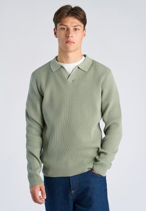 Giovane uomo con capelli castani corti che indossa un maglione verde chiaro a coste sopra una camicia bianca, con una mano nella tasca dei jeans blu.