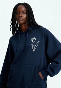 Sweat à capuche bleu marine avec une poche frontale, orné d'une broderie de tulipe blanche sur le côté gauche. Confectionné en tissu doux et texturé.