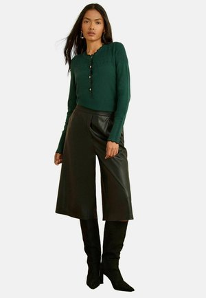Green knitted sweater with scalloped neckline, buttons down front; paired with black faux leather wide-leg trousers and knee-high boots.