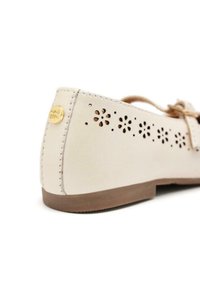 Chaussure en cuir beige clair avec des perforations florales sur le côté, un bout rond et une semelle plate en caoutchouc marron. Accent en logo doré à l'arrière.