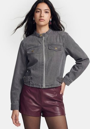 Young woman with long dark hair wears a gray zip-up jacket, burgundy leather shorts, black tights, and star-shaped earrings against a white background.