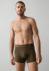 Barna boxeralsó puha textúrával, a deréknál logós pánttal. A modell semleges háttér előtt áll.