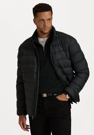 Man with freckles wearing a black puffer jacket over a black quarter-zip sweater, black pants, and a wristwatch, standing with one hand in pocket.