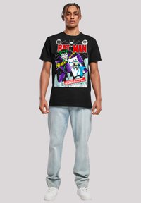 Black cotton T-shirt featuring comic art with Joker and Batman. Graphic includes bright colors, text, and a distinctive design. Worn with light jeans.