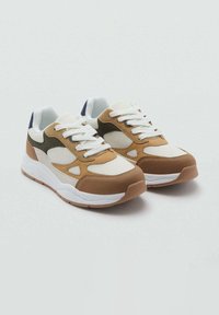 Kraftne športne čevlje z belim mrežastim materiali in tan, rjavimi ter črnimi usnjenimi plastmi, belimi vezalkami in debelimi belimi podplati na svetlo sivi podlagi.