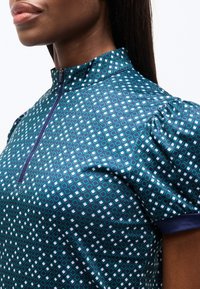 Γυναίκα που φοράει μπλε μπλούζα με σχέδια, ψηλό γιακά, κοντά φουσκωτά μανίκια και φερμουάρ μπροστά, σε απλό λευκό φόντο.