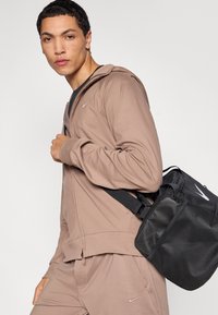 Ανοιχτό καφέ μπουφάν με φερμουάρ και κουκούλα, με λογότυπο Nike. Μαύρη αθλητική τσάντα με διακοσμητικά από δίχτυ και ιμάντα, τοποθετημένη στον ώμο του ατόμου.