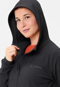 Felpa zip nera con superficie testurizzata, dotata di cappuccio e logo ricamato. Indossata sopra una maglietta arancione, con le mani che stringono la zip.