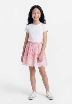 Mergina su ilgais juodais plaukais šypsosi, vilki baltą marškinėlių, rausvai baltą dryžuotą sijoną su nėrinių apdaila ir baltus sportinius batelius, stovėdama ant balto fono.