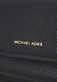 Juodas tekstūruotas odinis rankinės modelis su akivaizdžiu auksiniu "MICHAEL KORS" logotipu priekyje. Savybės apima elegantišką dizainą ir horizontalų siūlės detalumą.