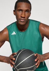 Zelený sieťový basketbalový dres s bielym logom Adidas, držaný hráčom, ktorý sa pevne drží čierneho basketbalu so striebornými akcentami.