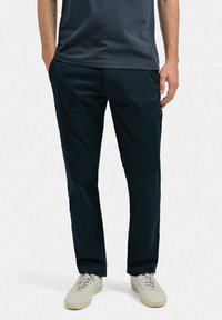Mörkblå chinos med en slät textur, rak bendesign, sidofickor och dragkedja framtill. Pairad med ljusa sneakers.