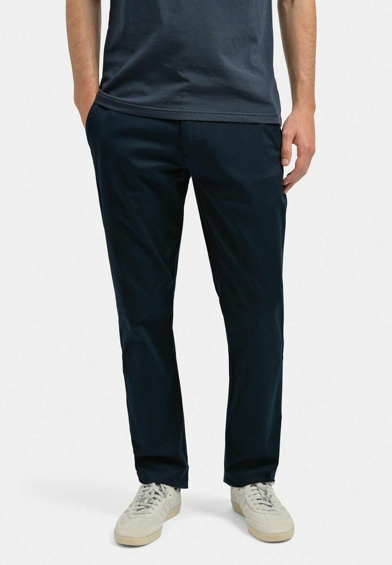 Mörkblå chinos med en slät textur, rak bendesign, sidofickor och dragkedja framtill. Pairad med ljusa sneakers.