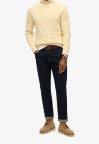 Pull à col roulé crème en point de câble, jean en denim foncé, ceinture en cuir marron et bottes à lacets couleur beige avec semelle beige. Look décontracté avec manches retroussées.