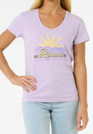 Frau trägt ein lavendelfarbenes T-Shirt mit V-Ausschnitt, bedruckt mit einer gelben Sonne und lila Wellen, sowie hellblaue Jeans, die Hand auf der Hüfte ruhend.