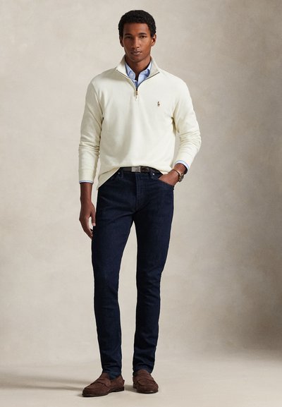 Άνδρας που φορά κρεμ πουλόβερ πάνω από μπλε πουκάμισο, σκούρο τζιν, καφέ μοκασίνια, στέκεται με το ένα χέρι στην τσέπη σε απλό φόντο.