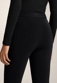 Zwarte legging gemaakt van een gladde, rekbare stof met een hoge tailleband. Heeft minimale branding aan de achterkant met een strakke uitstraling.