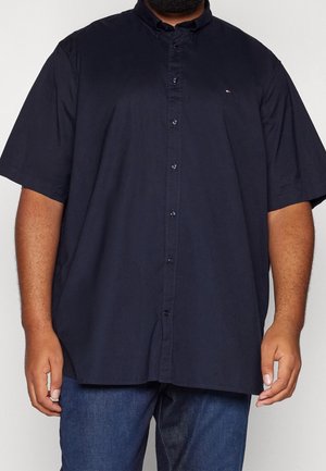 Mężczyzna w granatowej koszuli z krótkim rękawem zapinanej na guziki i niebieskich dżinsach, stojący na jednolitym jasnym tle.