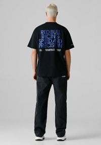 Person med kort blondt hår iført en sort Red Bull Racing t-shirt med blå tekst, sorte jeans og hvide sneakers, set bagfra.