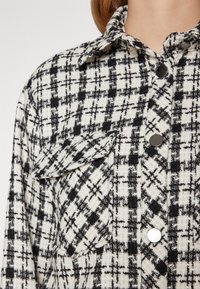 Ternede stofskjorte i sort og hvid, med krave, stor påsyet lomme og sølvknapdetaljer. Tydelig struktureret vævning.