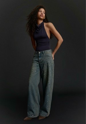 Vrouw met lang, krullend haar, gekleed in een mouwloze zwarte top en wijd uitlopende spijkerbroeken versierd met kleine studs, staande tegen een donkere achtergrond.