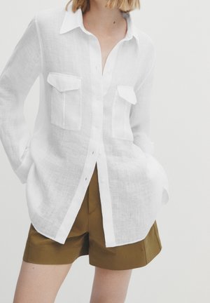 Woman wearing a loose white button-up shirt with chest pockets and brown knee-length shorts, hands in pockets, against a plain background.