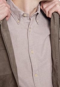 Vaaleanbeige painettavalla paidalla, jossa on kaulus ja beigejä nappeja, kerroksittain harmaan nahkatakin alla, jossa on vetoketjukiinnitys.