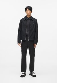 Melnā zip-up jaka ar studenēm apkaklē, kombinēta ar melnu pogu krekli un melnām biksēm. Apaviem ir melnbalti akcenti.