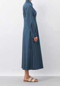 Abito lungo con colletto in tessuto blu intenso, caratterizzato da dettagli con bottoni sui polsini e una silhouette fluida, abbinato a sandali chiari.