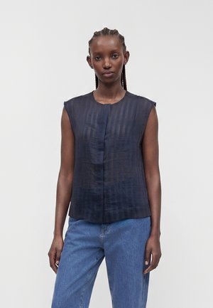 Femme aux cheveux tressés portant un haut noir transparent sans manches et un jean bleu, debout devant un fond clair uni.