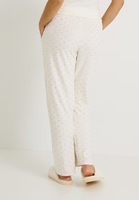 Pantalones de pijama crema claro con pequeños lunares dorados, con un corte relajado y cintura elástica. Combinados con zapatillas de casa crema y suaves.