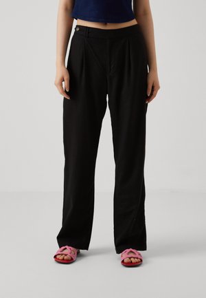 Pantalones de pierna ancha negros con cintura alta, frente con pliegues y cierre de botón. Llevados con sandalias rosa y rojas en forma de corazón.