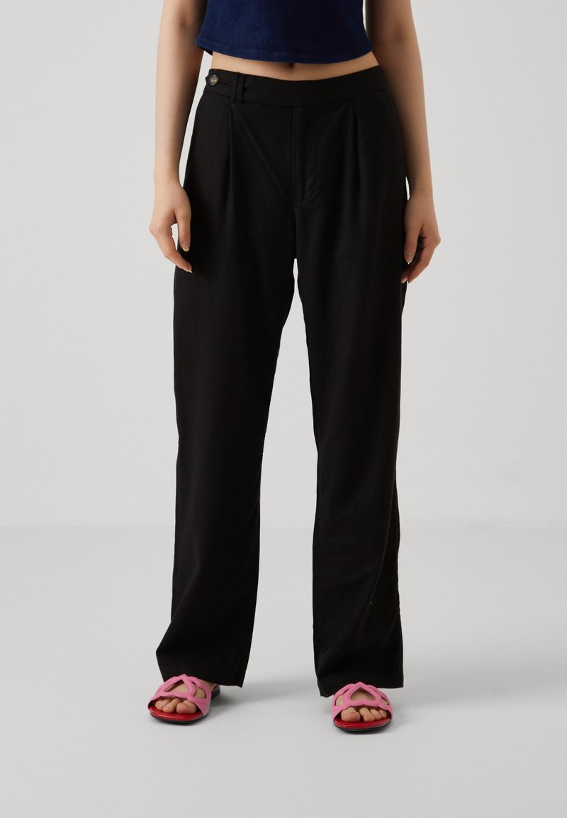 Pantalons larges noirs à taille haute, avec un devant plissé et une fermeture à bouton. Portés avec des sandales en forme de cœur rose et rouge.