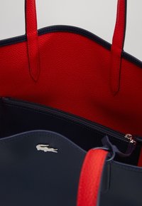 Sininen ja punainen kassi, jossa on teksturoitu ulkomateriaali, kaksoiskahvat ja vetoketjulla varustettu sisätasku. Pieni hopeinen logo yksityiskohtana vuorissa.