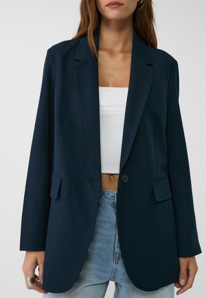 Femme portant un blazer oversize bleu foncé sur un crop top blanc et un jean bleu clair sur fond uni.