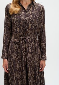 Sukienka koszulowa z długim rękawem w ciemnym wzorze węża. Wykonana z gładkiego materiału, z paskiem w talii i zapięciem na guziki, posiada dwie kieszenie na piersi.
