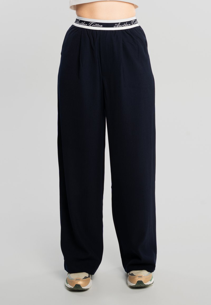 Pantalon large bleu marine avec taille élastique portant l'étiquette "Another Cotton", porté avec des baskets beige et blanc.