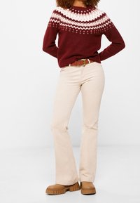 Pletený vínový svetr s bílým fair isle vzorem, doplněný béžovými rozšířenými kalhotami, hnědými botami na robustním podpatku a sladěným opaskem.