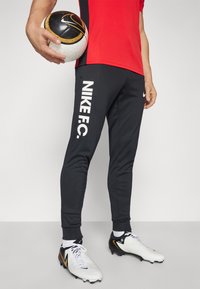 Schwarze Nike FC-Hosen mit weißem Branding, kombiniert mit weißen Fußballschuhen mit gelben Akzenten. Das Modell hält einen gemusterten Fußball.
