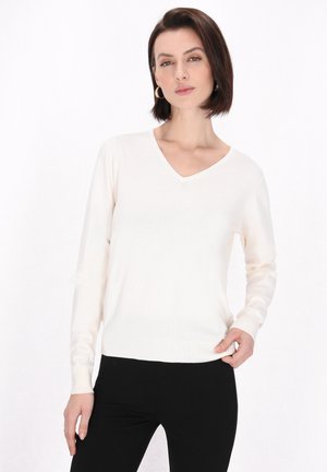 Vaalea beige V-pääntie neule, jossa on pitkät hihat, valmistettu pehmeästä kankaasta, jossa on raidoitetut hihansuut ja helma, päällä mustat housut.
