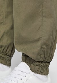 Pantalons oliviers fuselés avec des poignets élastiques, fabriqués en tissu lisse. Associés à des baskets blanches et des chaussettes blanches visibles.