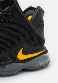 Scarpa sportiva nera con parte superiore in rete texturizzata, dettagli lucidi e un logo Nike giallo. Presenta una suola in gomma grigia con elementi trasparenti.