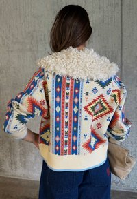 Γυναίκα με μακριά καστανά μαλλιά φοράει πολύχρωμο σακάκι με μεγάλο λευκό μάλλινο γιακά, στέκεται μπροστά σε τσιμεντένιο τοίχο, κρατώντας μια μπεζ τσάντα χειρός.