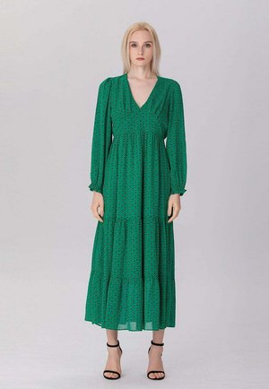 Femme blonde debout, portant une longue robe verte à pois noirs et des talons hauts noirs avec bride à la cheville, expression neutre, fond clair.