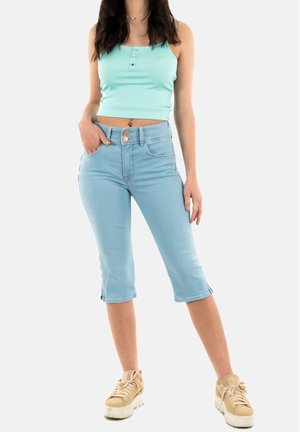 Femme portant un débardeur court bleu clair, un short en jean bleu clair au genou et des baskets beiges, debout avec une main dans la poche.
