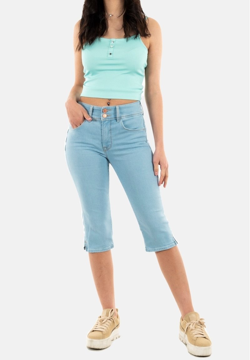 Femme portant un débardeur court bleu clair, un short en jean bleu clair au genou et des baskets beiges, debout avec une main dans la poche.