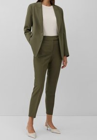 Oliwkowo zielony garnitur na miarę, z dopasowaną marynarką z wyciętymi klapami, wąskimi spodniami i kremowym, teksturowanym topem. Do tego jasnobeżowe szpilki.